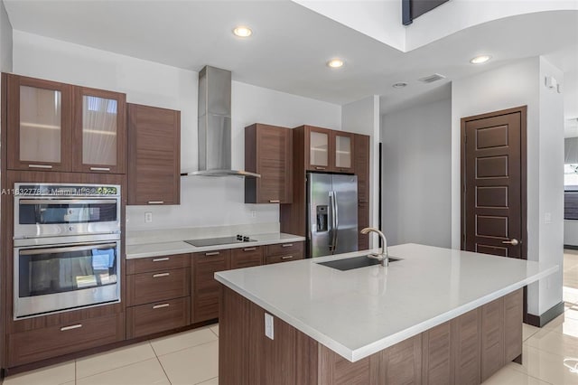 kitchen featuring light tile patterned flooring, wall chimney range hood, an island with sink, stainless steel appliances, and sink