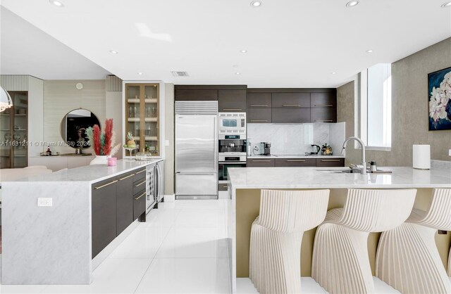 kitchen with light tile patterned flooring, kitchen peninsula, built in appliances, a kitchen bar, and sink