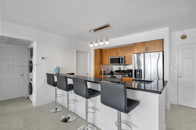 kitchen with stainless steel appliances, decorative light fixtures, light tile patterned floors, stacked washer and clothes dryer, and a kitchen breakfast bar