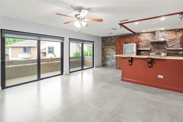 kitchen featuring light tile patterned flooring, ceiling fan, wall chimney range hood, stainless steel refrigerator, and a textured ceiling