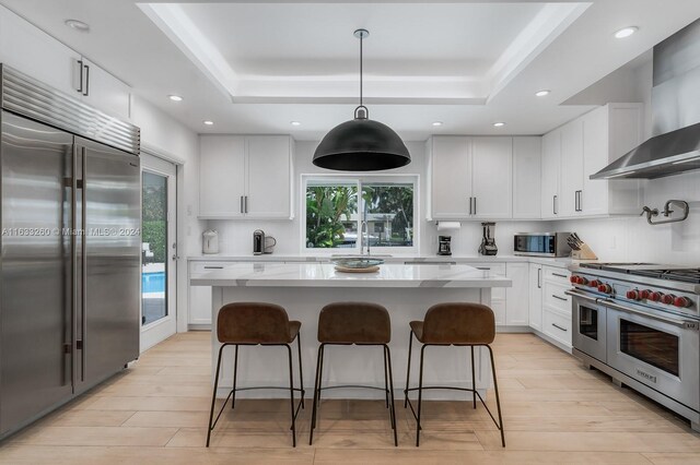 kitchen with high quality appliances, light hardwood / wood-style flooring, a tray ceiling, and wall chimney exhaust hood