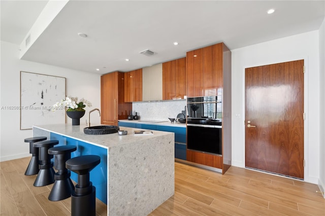 kitchen featuring oven, tasteful backsplash, an island with sink, a breakfast bar, and light hardwood / wood-style flooring