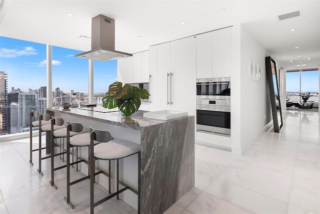 kitchen with light tile patterned flooring, white cabinets, a wall of windows, stainless steel double oven, and island range hood