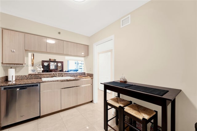 kitchen featuring dark stone counters, stainless steel dishwasher, light brown cabinetry, sink, and light tile patterned floors