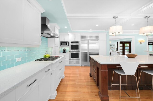 kitchen featuring wall chimney exhaust hood, pendant lighting, built in appliances, a center island with sink, and white cabinets