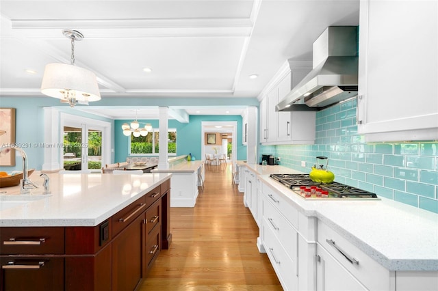 kitchen with light wood-type flooring, wall chimney exhaust hood, pendant lighting, white cabinetry, and an island with sink