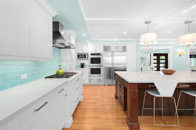 kitchen featuring wall chimney exhaust hood, built in appliances, light countertops, and a breakfast bar area