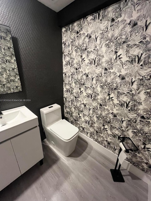 half bathroom with toilet, wallpapered walls, wood finished floors, and vanity