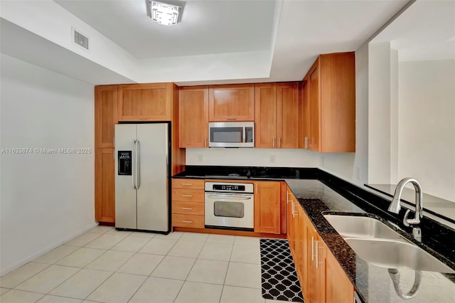 kitchen with appliances with stainless steel finishes, a sink, dark stone counters, a raised ceiling, and brown cabinetry