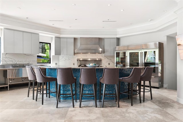 kitchen featuring built in refrigerator, wall chimney range hood, gray cabinets, a kitchen bar, and decorative backsplash