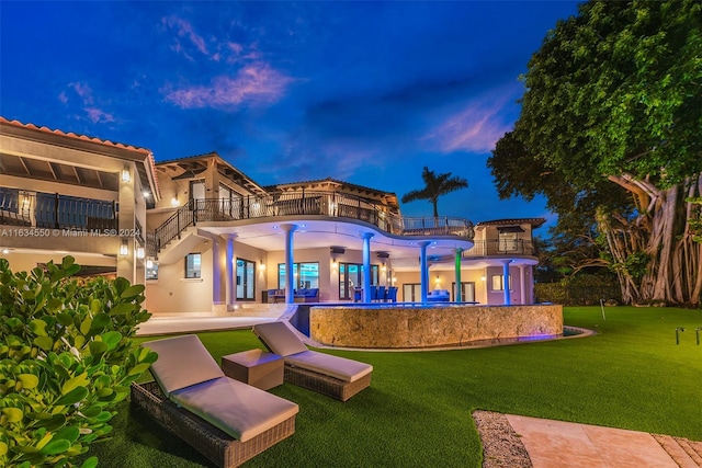 back house at dusk featuring a balcony, a swimming pool, a lawn, and a patio