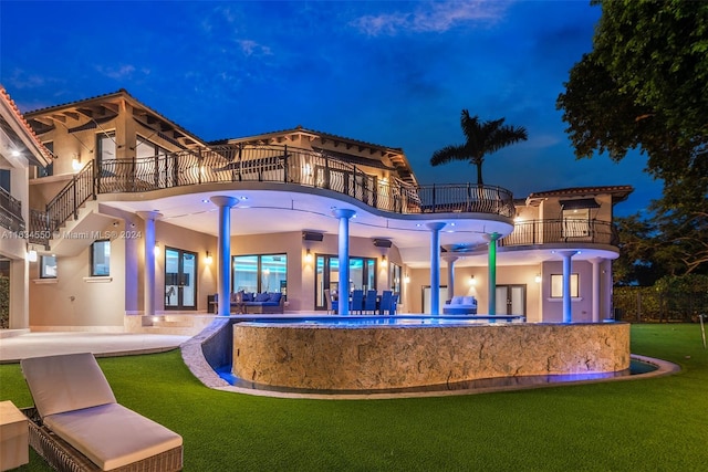 back house at night featuring a balcony, a patio area, and a lawn