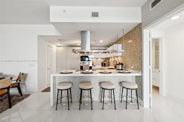 kitchen with backsplash, kitchen peninsula, double oven, island range hood, and a breakfast bar