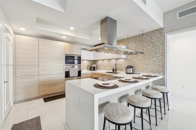 kitchen with island exhaust hood, a tray ceiling, black appliances, light brown cabinets, and a breakfast bar area