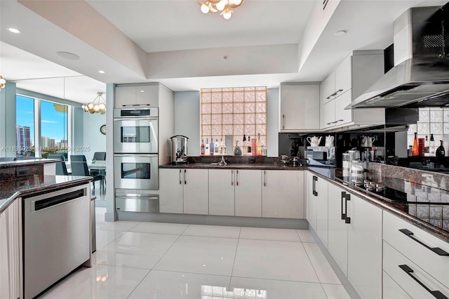 kitchen featuring a warming drawer, an inviting chandelier, stainless steel appliances, a raised ceiling, and wall chimney range hood