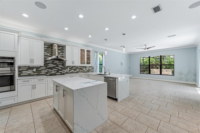 kitchen with backsplash, a kitchen island, ceiling fan, appliances with stainless steel finishes, and wall chimney exhaust hood