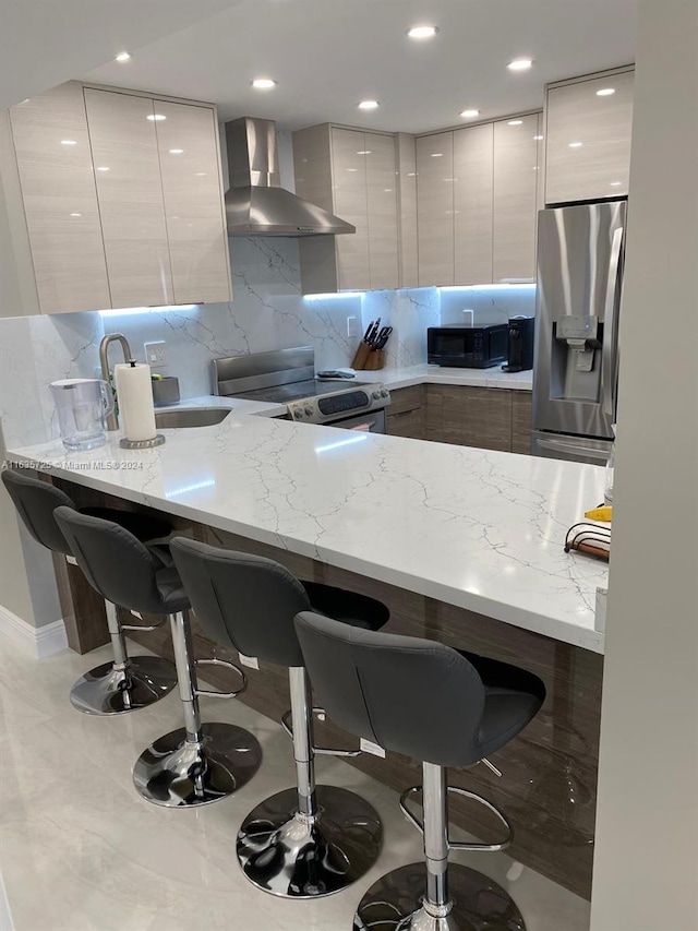 kitchen with a kitchen bar, light stone counters, wall chimney range hood, and appliances with stainless steel finishes