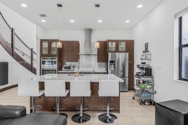 kitchen featuring a kitchen island with sink, wall chimney exhaust hood, appliances with stainless steel finishes, decorative backsplash, and decorative light fixtures