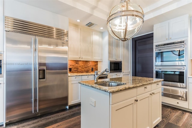 kitchen featuring white cabinetry, sink, hanging light fixtures, built in appliances, and a kitchen island with sink