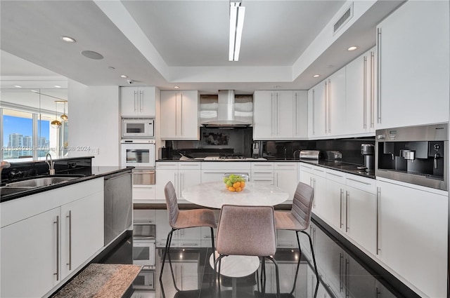 kitchen with white appliances, backsplash, white cabinetry, a raised ceiling, and wall chimney range hood