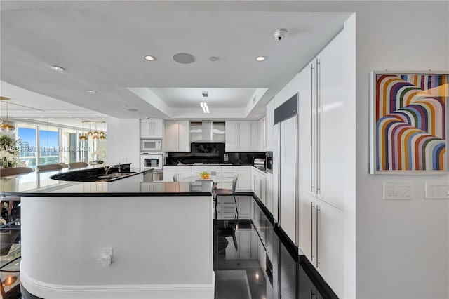 kitchen featuring tasteful backsplash, white appliances, a tray ceiling, white cabinets, and wall chimney range hood