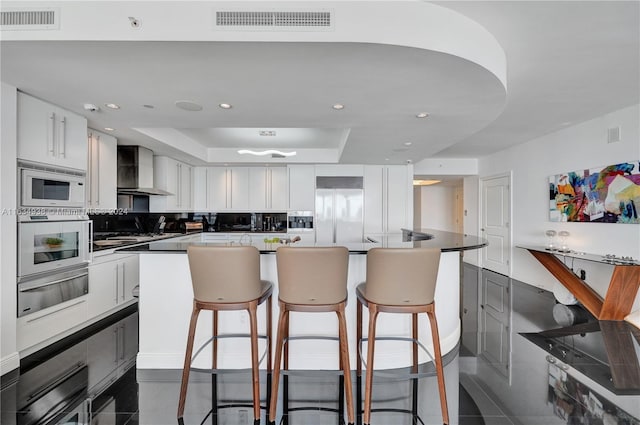 kitchen featuring dark tile patterned floors, appliances with stainless steel finishes, white cabinetry, wall chimney range hood, and a tray ceiling