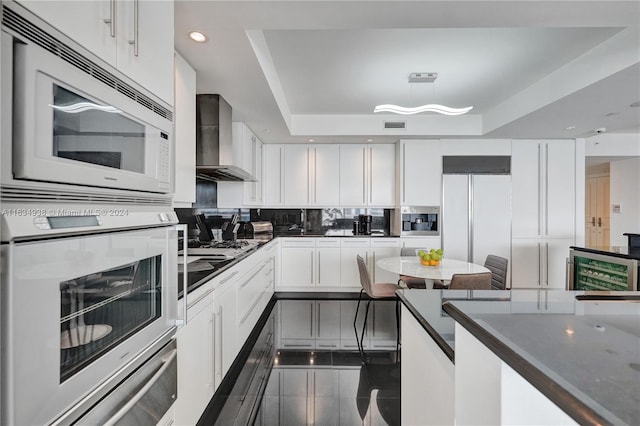 kitchen featuring built in appliances, white cabinetry, wall chimney exhaust hood, a tray ceiling, and dark countertops