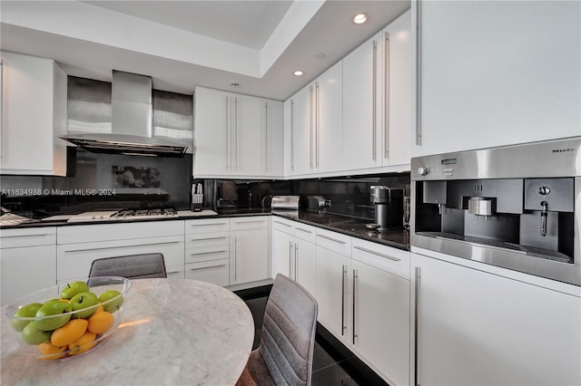 kitchen featuring tasteful backsplash, a raised ceiling, tile patterned floors, white cabinets, and wall chimney range hood