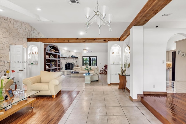 living room with ceiling fan with notable chandelier, light hardwood / wood-style floors, a stone fireplace, and beam ceiling