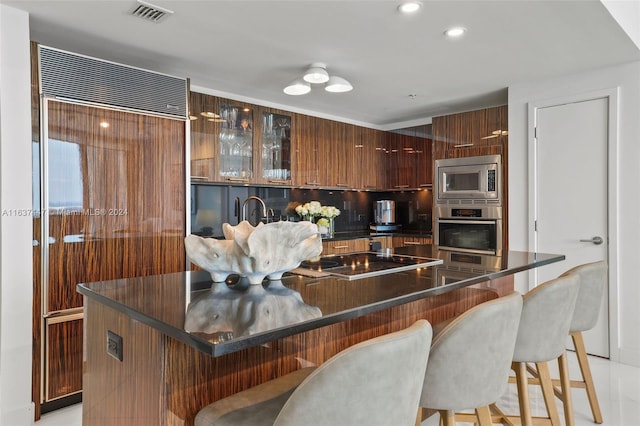 kitchen with light tile patterned floors, dark brown cabinets, a breakfast bar, built in appliances, and tasteful backsplash