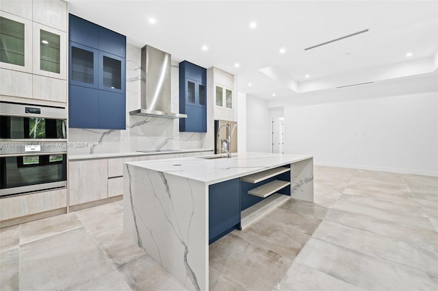 kitchen with light stone countertops, a center island with sink, double oven, a raised ceiling, and wall chimney exhaust hood