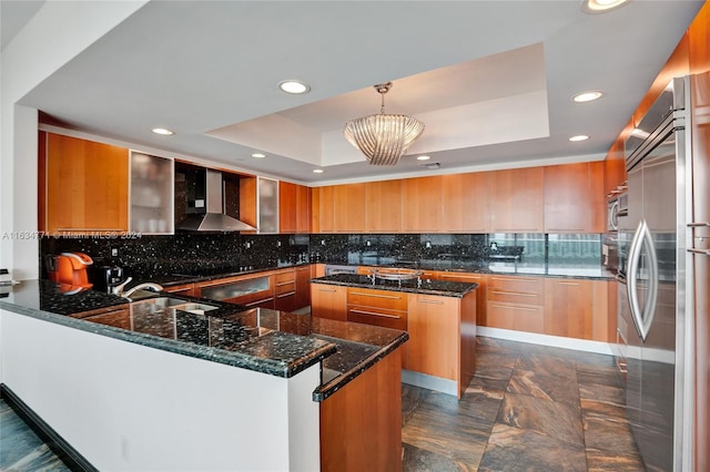 kitchen with wall chimney range hood, a raised ceiling, appliances with stainless steel finishes, a center island, and decorative light fixtures