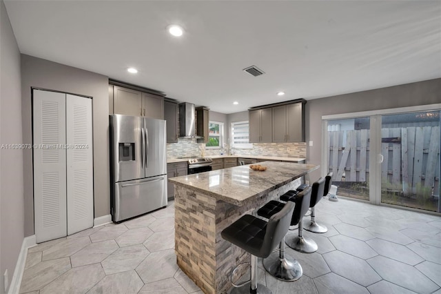 kitchen featuring appliances with stainless steel finishes, light stone countertops, wall chimney range hood, a kitchen breakfast bar, and a kitchen island