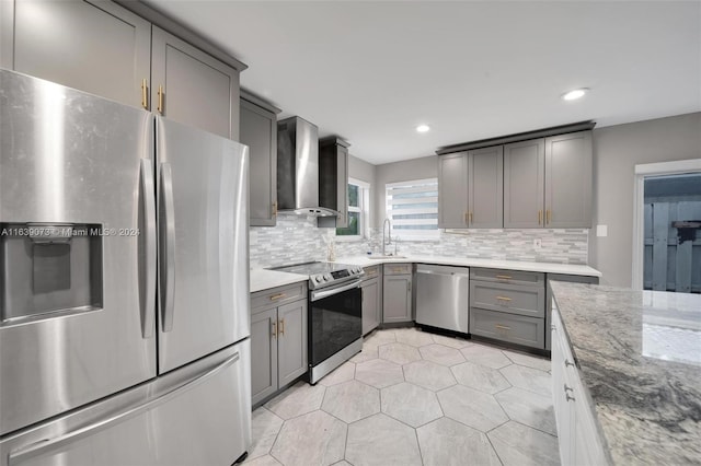 kitchen featuring gray cabinetry, appliances with stainless steel finishes, light stone counters, wall chimney exhaust hood, and tasteful backsplash