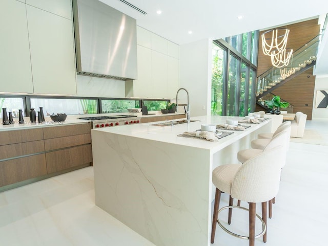 kitchen featuring sink, a kitchen island with sink, wall chimney range hood, white cabinetry, and a breakfast bar area