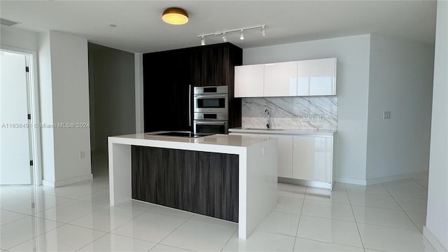 kitchen with backsplash, white cabinetry, double oven, and a kitchen island with sink