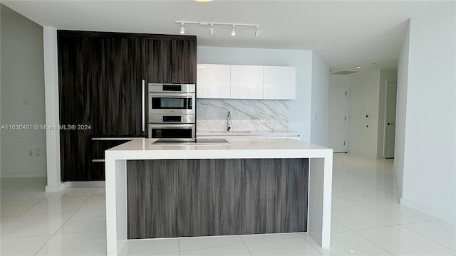 kitchen featuring a kitchen island with sink, white cabinets, decorative backsplash, dark brown cabinets, and stainless steel double oven