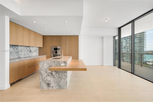 kitchen featuring expansive windows, decorative backsplash, oven, sink, and a kitchen island with sink