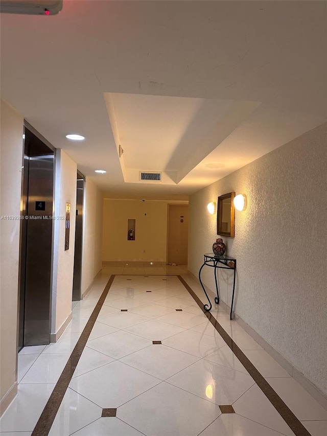 corridor with elevator, light tile patterned flooring, and a raised ceiling