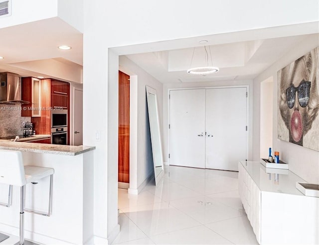 kitchen featuring wall chimney range hood, light tile patterned floors, double oven, decorative backsplash, and decorative light fixtures