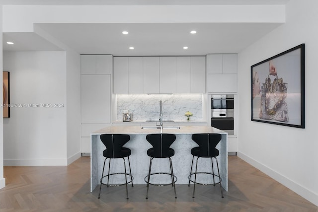 kitchen with white cabinetry, double oven, a center island with sink, parquet flooring, and sink