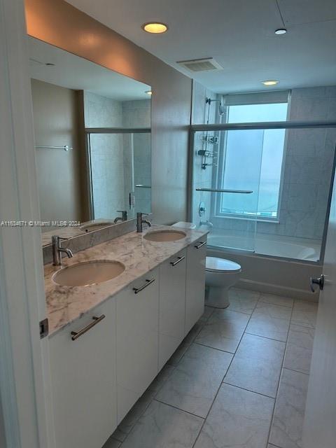 full bathroom featuring combined bath / shower with glass door, dual vanity, toilet, and tile patterned flooring