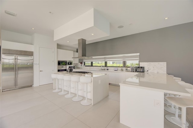 kitchen with white cabinetry, built in appliances, a breakfast bar area, and wall chimney range hood