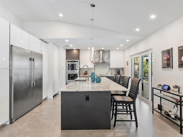 kitchen with white cabinets, light stone counters, vaulted ceiling, stainless steel appliances, and wall chimney range hood