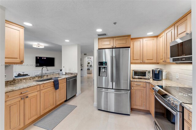 kitchen featuring stainless steel appliances, sink, light brown cabinetry, light stone counters, and a textured ceiling