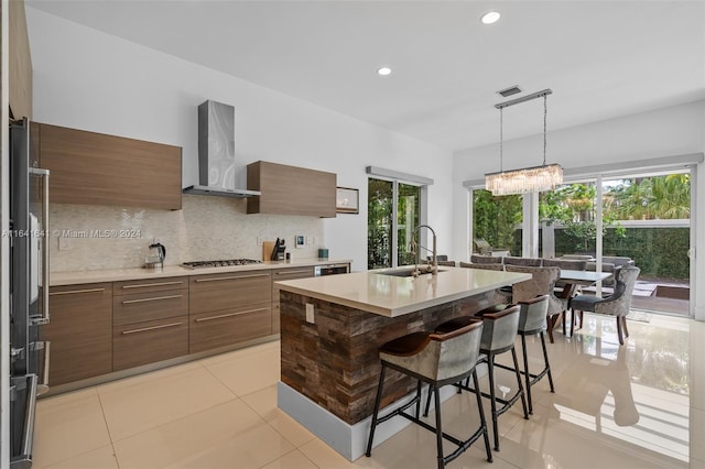 kitchen featuring wall chimney exhaust hood, tasteful backsplash, sink, a center island with sink, and light tile patterned floors