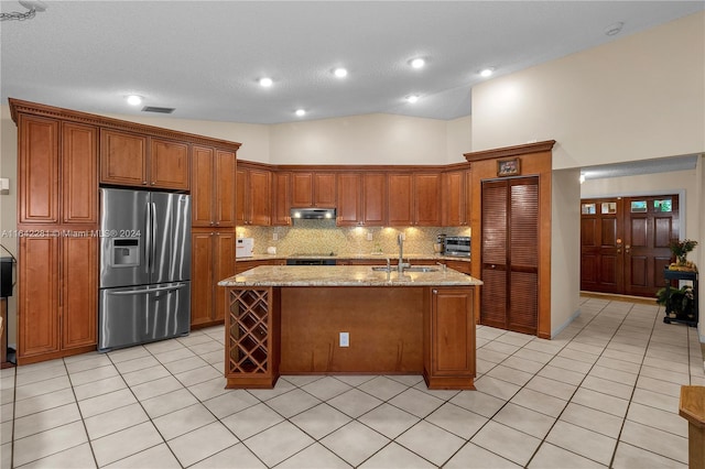 kitchen with stainless steel fridge, light stone counters, sink, a center island with sink, and high vaulted ceiling