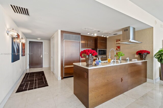 kitchen featuring wall chimney range hood, built in appliances, track lighting, light tile patterned floors, and kitchen peninsula