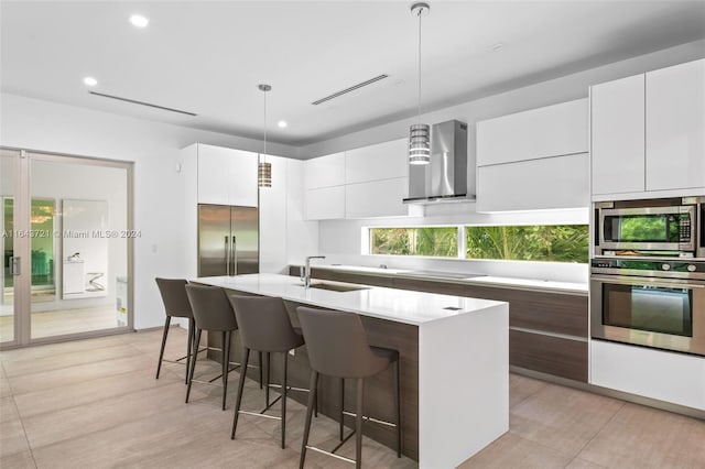 kitchen featuring decorative light fixtures, an island with sink, built in appliances, wall chimney range hood, and white cabinets