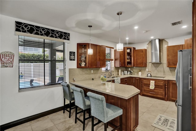 kitchen featuring kitchen peninsula, wall chimney exhaust hood, stainless steel refrigerator, hanging light fixtures, and a breakfast bar area